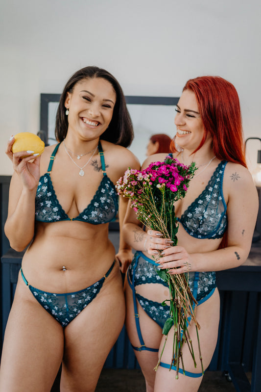 Two women in matching blue and green bikinis holding flowers and a fruit.
