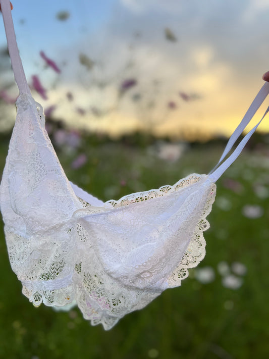 A white lace bralette with adjustable straps, displayed against an outdoor backdrop.