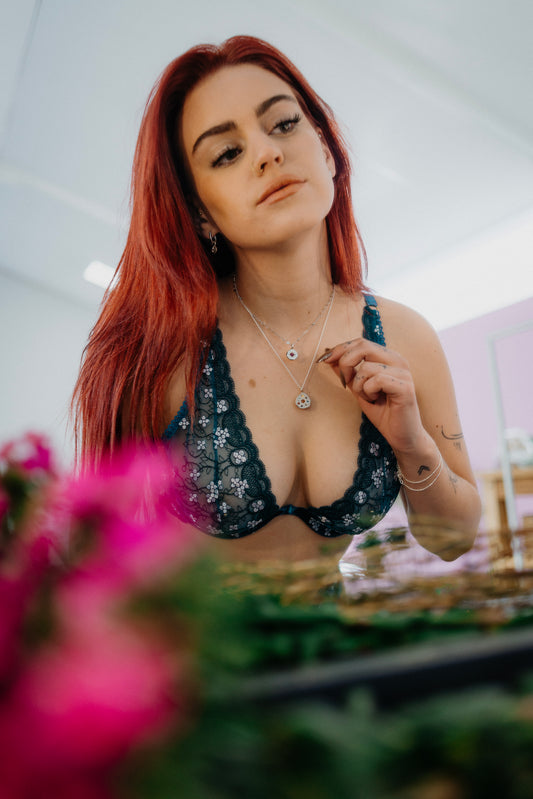 A woman wearing a Jade bra with turquoise green lace and white floral accents, with underwire and gold hardware, standing in front of a pink flower arrangement.
