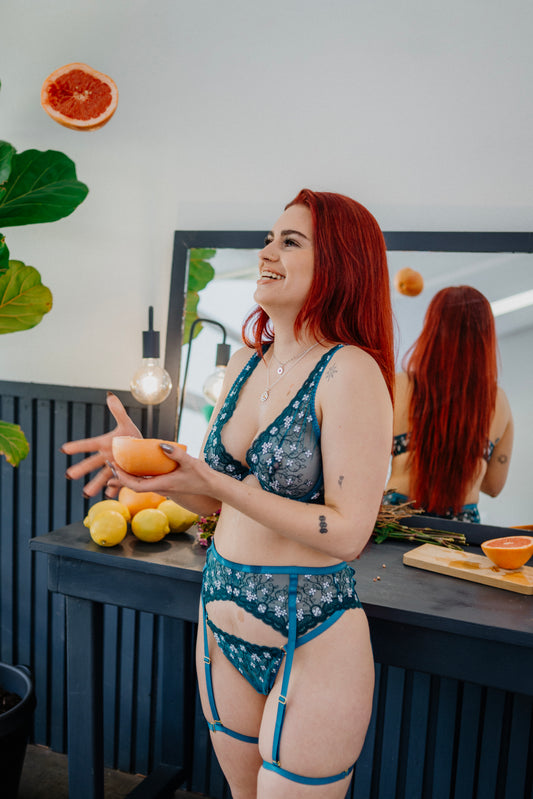 This image features a woman modeling the Jade G-String, which is presented against a backdrop of a modern kitchen setting. The woman is wearing the G-string, which is prominently displayed, along with a few other pieces of intimate apparel, such as a bra and a garter belt, which are also visible in the background.