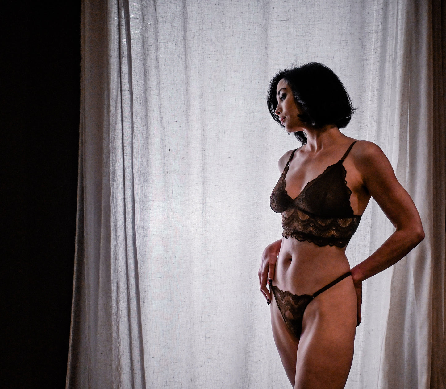 Woman wearing a mocha brown lace lingerie set against a white curtain.